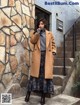 A woman wearing a brown coat standing in front of a stone wall.