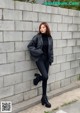 A woman leaning against a wall wearing a black jacket and black pants.