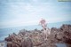 A woman in a bikini and hat standing on rocks by the ocean.